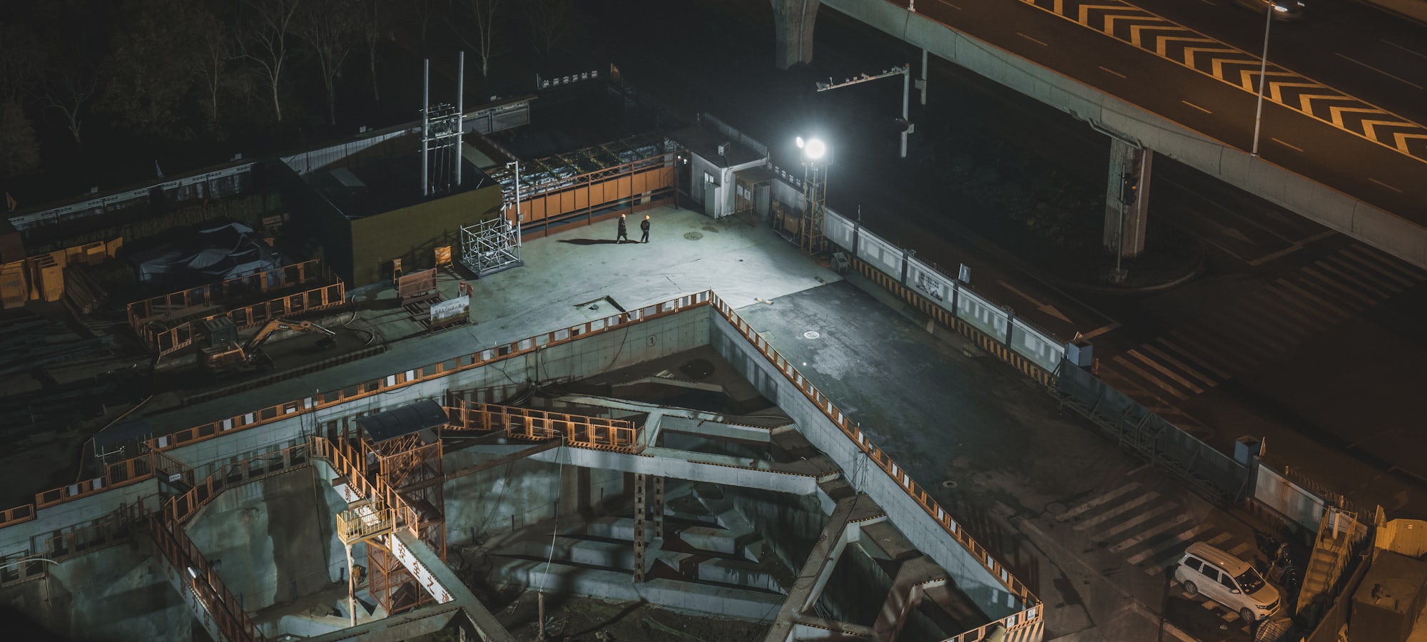 An aerial view of a lit construction site alongside an elevated roadway at night
