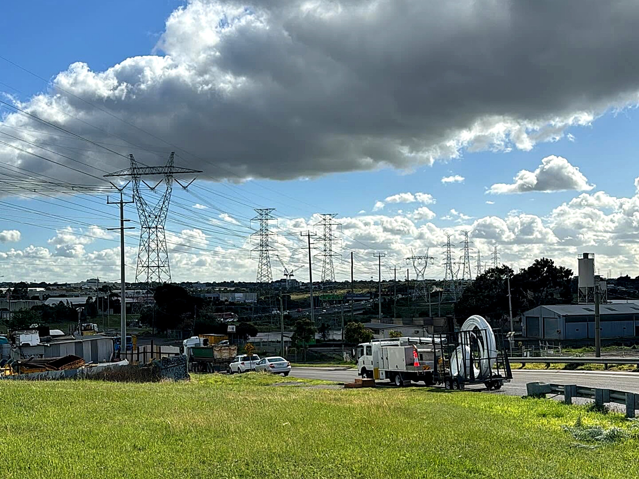 An All Network Connections service truck and a trailer of coiled conduit on a freeway verge, transmission towers and industrial sheds behind