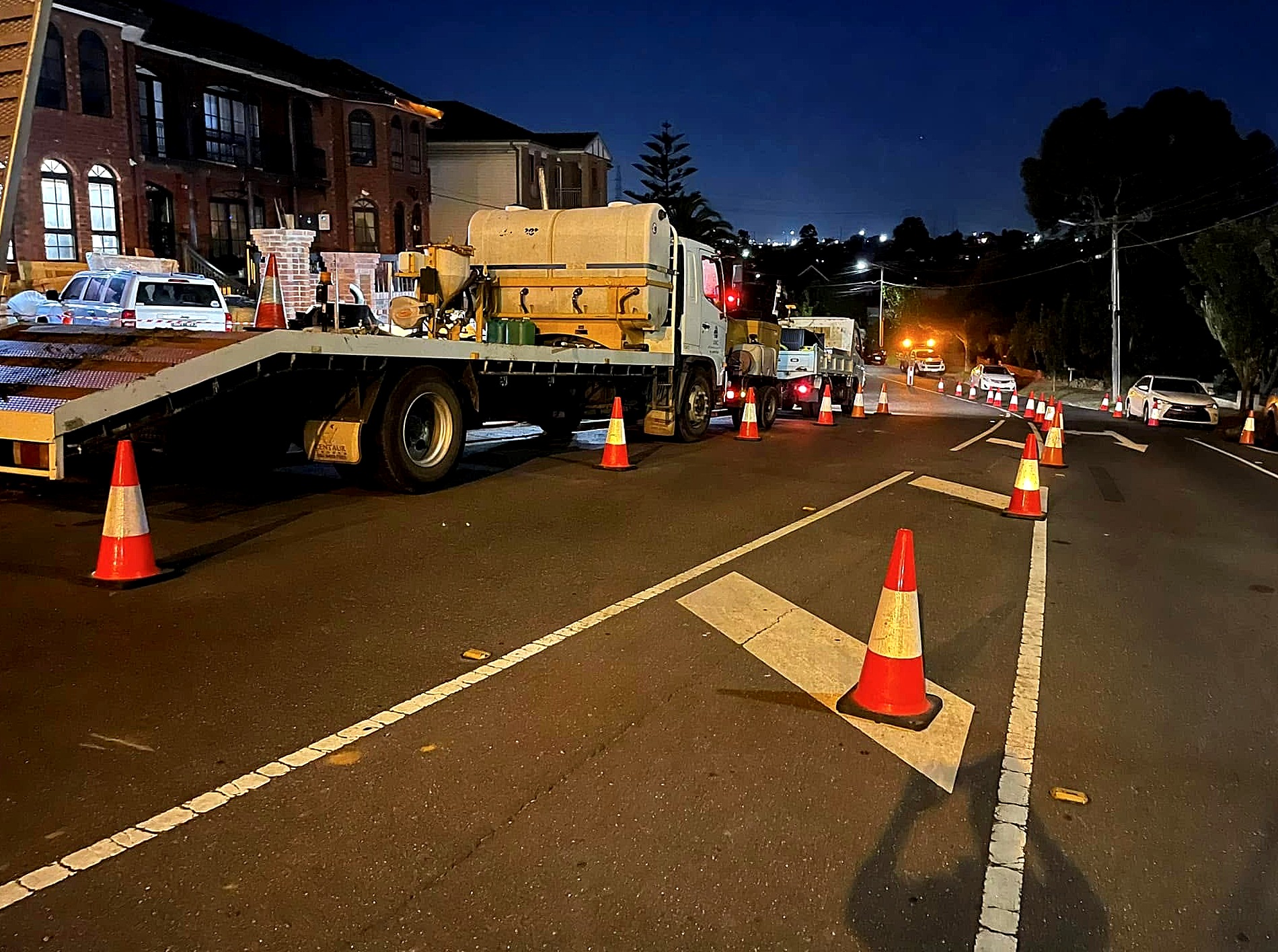 A night road job under traffic management — the All Network Connections float and tipper on a coned-off lane, with the cone taper running away down the carriageway